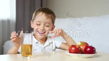 一个小男孩坐在桌子旁。 桌子上有一杯果汁和苹果。 孩子喝了一杯果汁